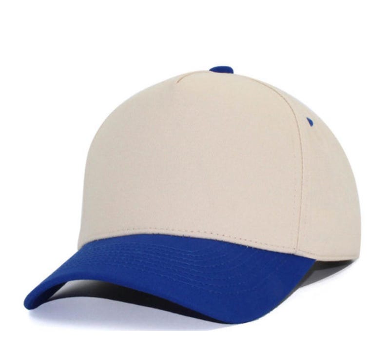 May include: A five-panel baseball cap featuring a solid cream-colored crown and a contrasting vibrant blue curved brim. The cap includes a small blue button at the top center and blue eyelets on the side panels, providing a clean and structured look.