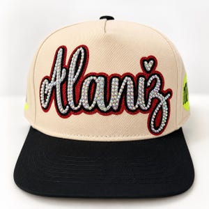 May include: A beige baseball cap with a black brim featuring the name Alaniz in cursive script. The lettering is outlined in red and filled with iridescent rhinestones, with a small rhinestone heart above the letter i. A neon yellow patch is visible.