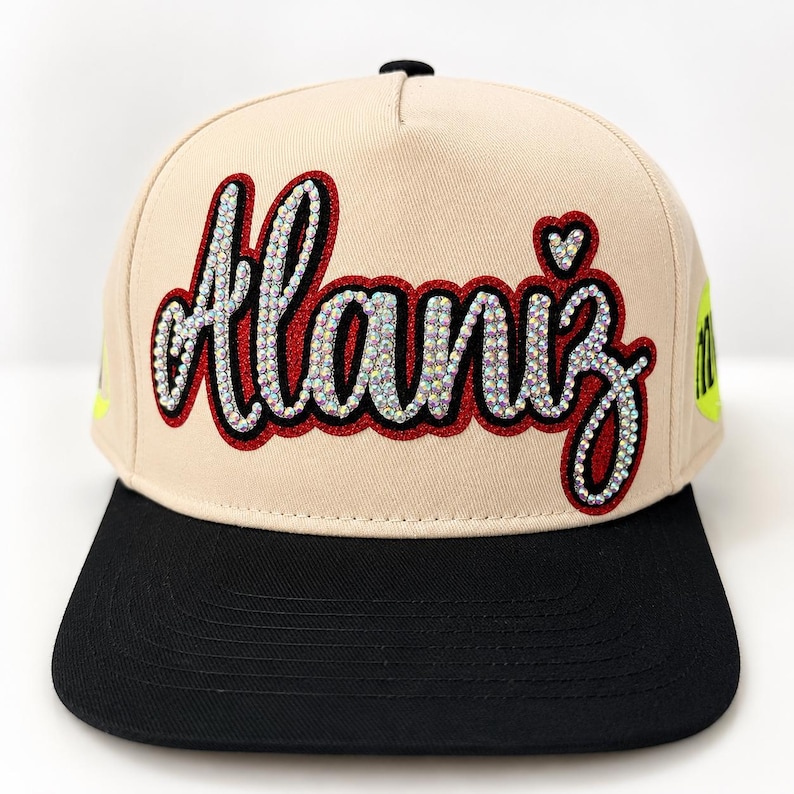 May include: A beige baseball cap with a flat black brim. The front features the name Alaniz in cursive script, embellished with iridescent rhinestones and outlined in dark red. A small rhinestone heart sits above the letter i.