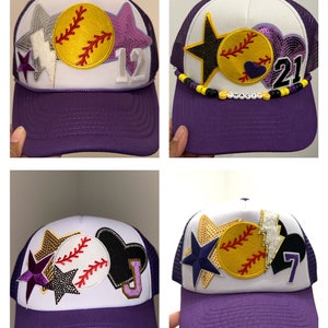 May include: Four purple and white baseball caps with different patches and embellishments. The caps have a mesh back and a white front with a variety of patches, including stars, hearts, lightning bolts, and a softball. The caps also have numbers, including 12, 21, 7, and a letter U.