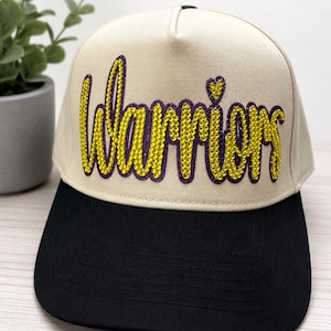 May include: A beige baseball cap with a black brim featuring the word Warriors in yellow rhinestones with a purple outline. The script lettering includes a small heart shape above the letter i. The cap has a structured crown and a flat brim.
