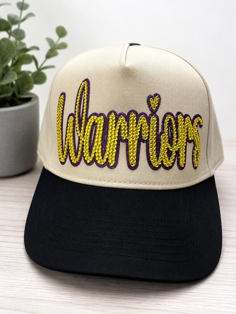 May include: A beige baseball cap with a black brim featuring the word Warriors in yellow rhinestones with a purple outline. The script lettering includes a small heart shape above the letter i. The cap has a structured crown and a flat brim.