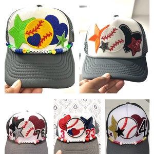 May include: Four gray and white baseball caps with different colored patches and embellishments. The caps have a mesh back and a gray brim. The patches include a baseball, a heart, a star, and numbers. The caps are decorated with beads and charms.