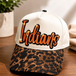 May include: A baseball cap with a white crown and a brown and black leopard print brim. The front of the crown features the word "Indians" in orange rhinestones with a black outline. The cap has a small black button on the top center.
