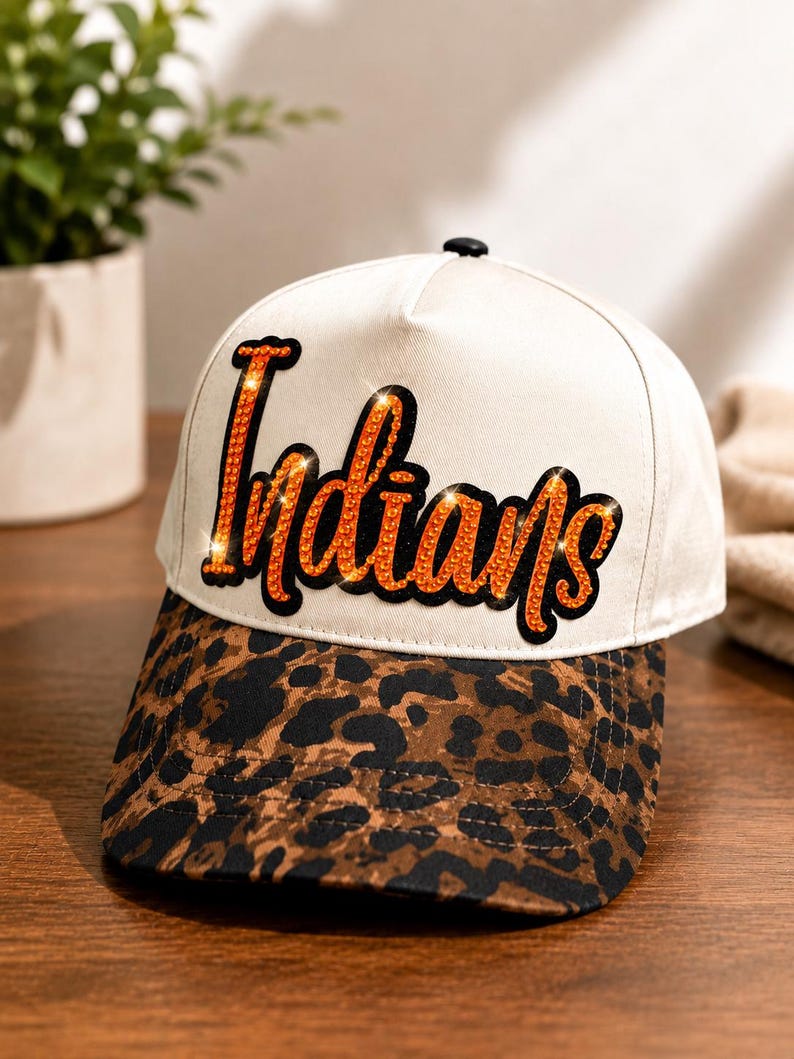 May include: A baseball cap with a white crown and a brown and black leopard print brim. The front of the crown features the word "Indians" in orange rhinestones with a black outline. The cap has a small black button on the top center.