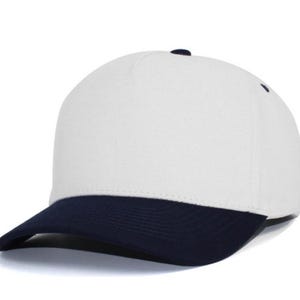 May include: A five-panel baseball cap featuring a solid white crown and a contrasting dark navy blue curved brim. The cap includes a small dark navy blue button at the top center and matching dark navy blue eyelets on the side panels for ventilation.