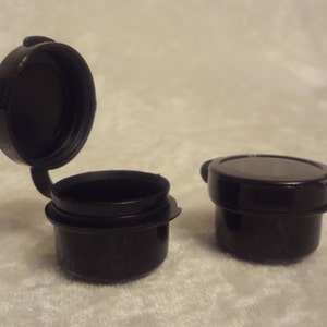 May include: Two small, black plastic containers with hinged lids. One lid is open, revealing the interior. The other container has its lid closed. The containers are on a light-colored surface.
