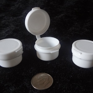 May include: Several small, white plastic containers with hinged lids are displayed. One lid is open, revealing the interior. A U.S. dime is included for size comparison. The containers are likely for small items or samples.