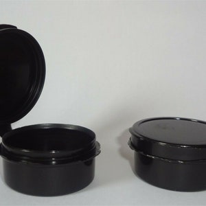 May include: Two small, black, round containers with hinged lids. One container's lid is open, revealing the interior. The other container's lid is closed. The containers have a simple, functional design.