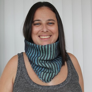 May include: A knitted cowl in shades of blue and teal, with vertical stripes. The cowl is worn around the neck and has a triangular shape at the front. The material appears soft and warm, suitable for cooler weather.
