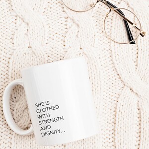 May include: White ceramic mug with a black inscription that reads "She is clothed with strength and dignity..." on a white knitted surface with a pair of gold-rimmed glasses.