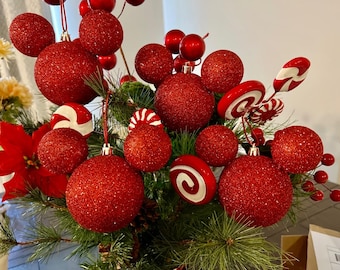 May include: A festive arrangement of red glitter ornaments, candy cane decorations, and berries nestled among evergreen branches. The ornaments vary in size and shape, creating a vibrant holiday display.