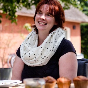 K&ouml;nnte beinhalten: Eine Frau mit roten Haaren tr&auml;gt einen wei&szlig;en Schal mit schwarzen Diamantenmustern. Sie l&auml;chelt und schaut nach oben. Sie tr&auml;gt ein schwarzes Oberteil und sitzt an einem Tisch mit einem Glas Eistee und Muffins.