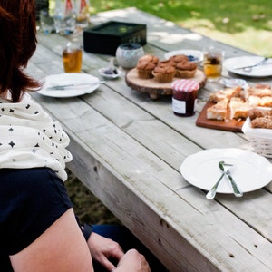 K&ouml;nnte beinhalten: Eine Frau sitzt an einem Holztisch im Freien und genie&szlig;t eine Auswahl an Geb&auml;ck, darunter Muffins und Croissants. Es gibt auch Teller mit Essen, Gl&auml;ser mit Getr&auml;nken und eine wei&szlig;e Keramiksch&uuml;ssel gef&uuml;llt mit Geb&auml;ck.