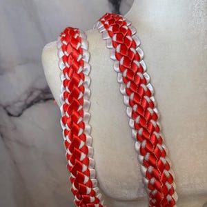 May include: A close-up of a braided lei made from red and white satin ribbons. The lei is displayed against a soft, off-white background, highlighting the detailed weaving. A gold trim adds a touch of elegance to the design.
