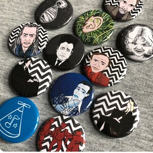 May include: A selection of round pin badges with colourful illustrations of characters and symbols. The badges feature portraits, a blue party hat, and a black and white zigzag pattern. The badges are arranged on a grey surface.