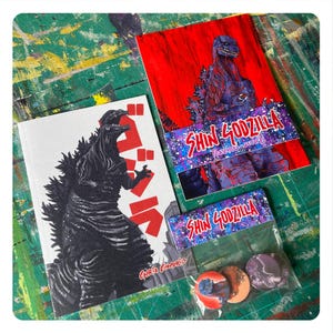 May include: A selection of Shin Godzilla merchandise, including a book with a black and white Godzilla illustration and Japanese text, postcards, and a set of three round badges. The postcards feature a red background and a purple Godzilla.