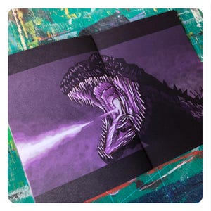 Op de afbeelding: Een paarse en zwarte illustratie van een monster met scherpe tanden en een gloeiende mond, gedrukt op de pagina's van een boek.