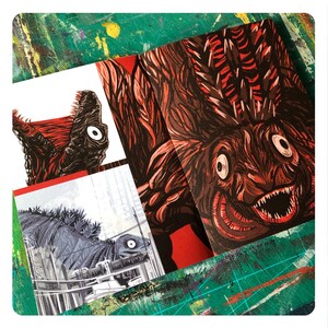 Op de afbeelding: Een close-up van een rode en zwarte monsterillustratie met scherpe tanden en grote ogen. De illustratie is gedrukt op een gevouwen kaart. De kaart ligt op een groen oppervlak.