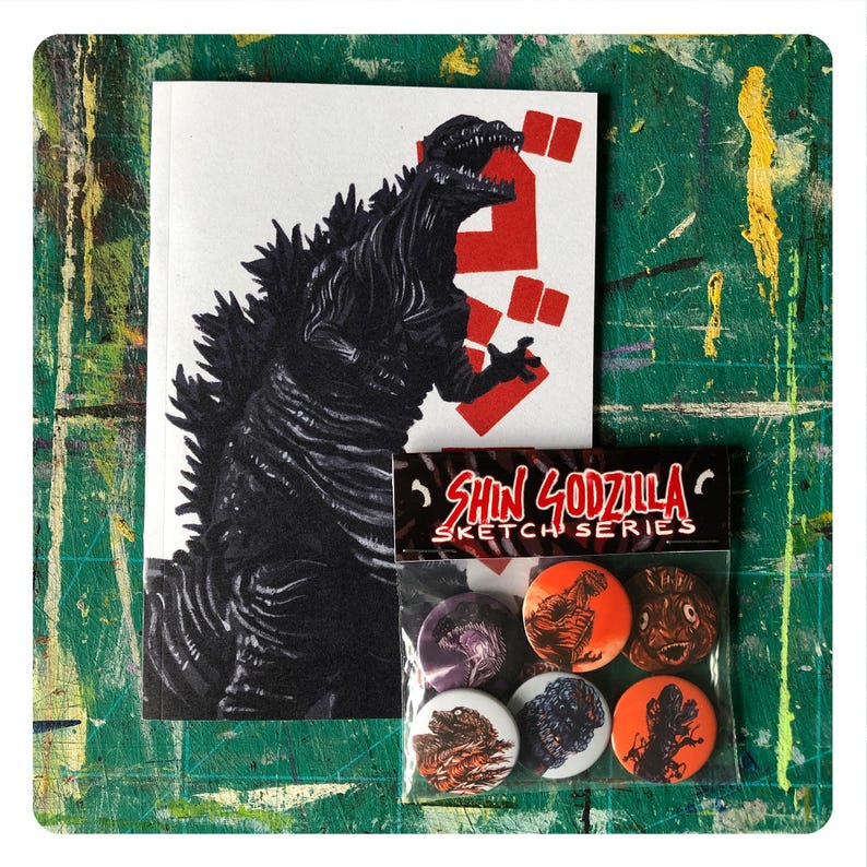 Op de afbeelding: Een zwart-witte illustratie van Godzilla op een witte kaart met rode Japanse karakters. Een plastic zakje met zes kleurrijke knopen met Godzilla in verschillende poses. Het zakje heeft de tekst "Shin Godzilla Sketch Series".