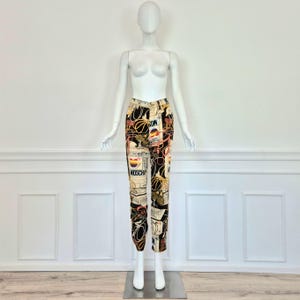 May include: Black and cream colored pants with a colorful print featuring the word "Moschino" and other graphic designs.