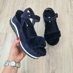 May include: Navy blue suede sandals with a fluffy, dark blue fur lining. The sandals have a thick sole with a white stripe and a buckle closure on the ankle strap. The shoes are displayed on a wooden surface.