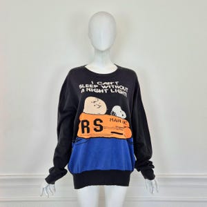 Peut inclure: Sweat-shirt noir avec un motif bleu et orange de Snoopy et Charlie Brown dans un radeau de sauvetage. Le texte sur le sweat-shirt est "I can't sleep without a night light!"