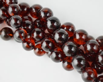 May include: A close-up of a strand of dark amber-colored, round beads. The beads have a glossy, reflective surface and are arranged diagonally across a white background. The beads are uniform in size and shape, suggesting they are for jewelry making.