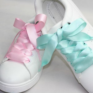 Op de afbeelding: Een paar witte sneakers met roze en lichtblauwe satijnen lintjes als veters. De veters zijn aan elke schoen in een strik geknoopt.