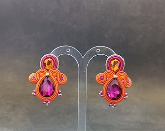May include: A pair of dangle earrings with orange, pink, and gold accents. The earrings feature a large, faceted, pink gemstone in the center.