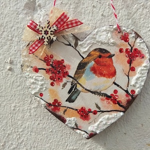 May include: A heart-shaped wooden ornament with a robin perched on a branch with red berries. The ornament is decorated with faux snow and a red and white gingham bow with a wooden snowflake.