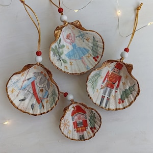Könnte beinhalten: Vier Weihnachtsornamente aus Muscheln mit goldenem Rand, die eine Ballerina und einen Nussknacker zeigen. Jedes Ornament hat eine goldene Aufhängeschnur mit roten und weißen Perlen.