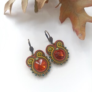Peut inclure: Boucles d'oreilles artisanales avec un grand cristal orange au centre, entouré de perles et de fils colorés rouges, verts et dorés. Les boucles d'oreilles ont une fermeture à levier. Le design évoque l'automne.