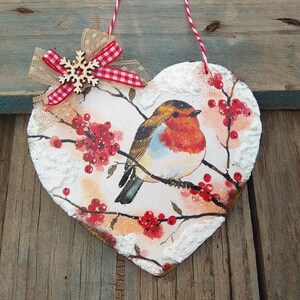 May include: A heart-shaped wooden ornament with a robin perched on a branch with red berries. The ornament is decorated with white paint to resemble snow and has a red and white gingham bow with a wooden snowflake.