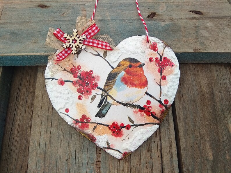 May include: A heart-shaped wooden ornament with a robin perched on a branch with red berries. The ornament is decorated with white paint to resemble snow and has a red and white gingham bow with a wooden snowflake.