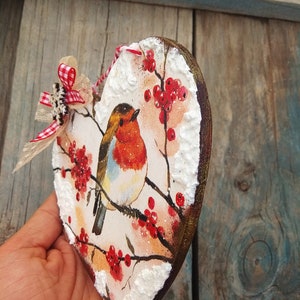 May include: A wooden heart-shaped ornament with a painted image of a robin perched on a branch with red berries. The background is white with a snowy effect.