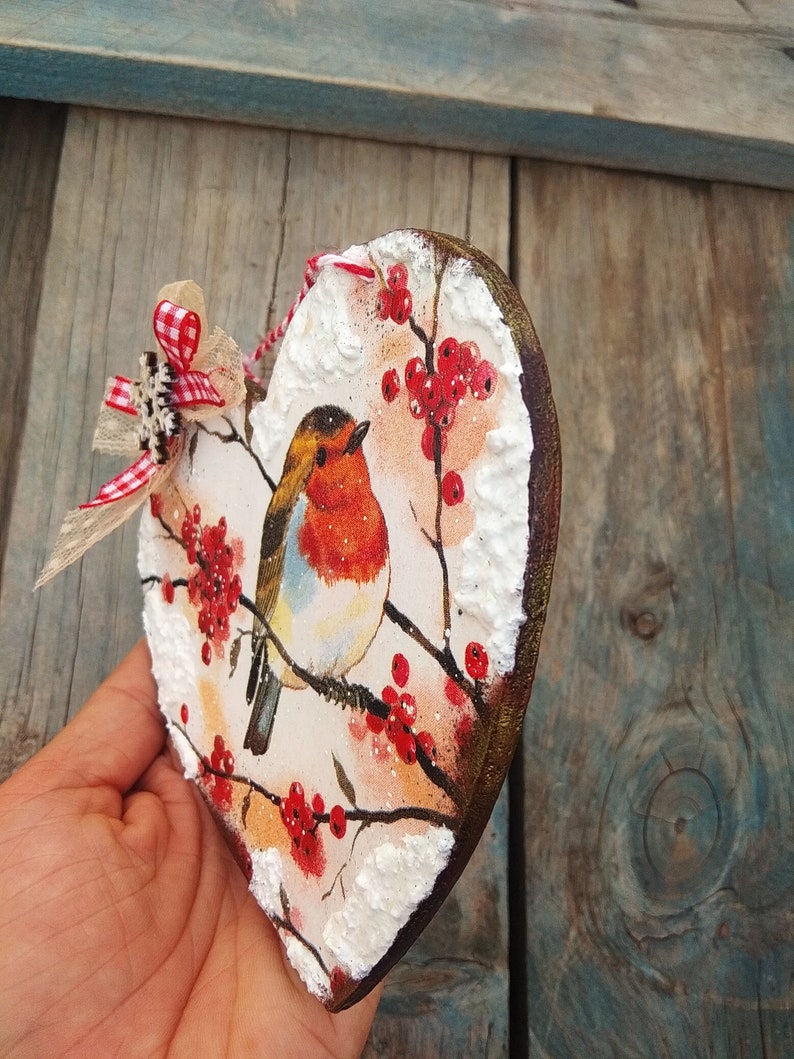 May include: A wooden heart-shaped ornament with a painted image of a robin perched on a branch with red berries. The background is white with a snowy effect.