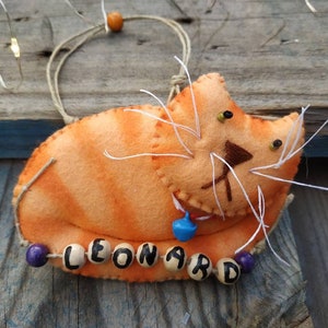 Könnte beinhalten: Orangefarbener Filzkatzen-Anhänger mit einer blauen Glocke und dem Namen "LEONARD", der mit Holzperlen ausgeschrieben ist.