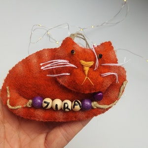 May include: Orange felt cat with embroidered white whiskers and yellow nose. The cat has purple and wooden beads with the name "JIRA" spelled out.