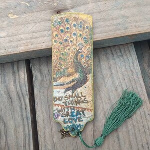 May include: A gold bookmark with a peacock design and the text "Do small things with great love" printed on it. The bookmark has a green tassel.