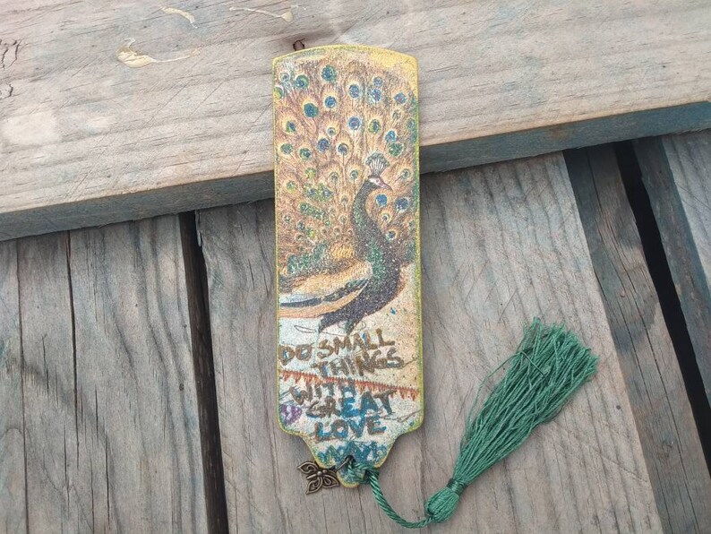 May include: A gold bookmark with a peacock design and the text "Do small things with great love" printed on it. The bookmark has a green tassel.
