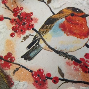 May include: A robin perched on a branch with red berries, painted on a heart-shaped wooden surface with a snowy texture.