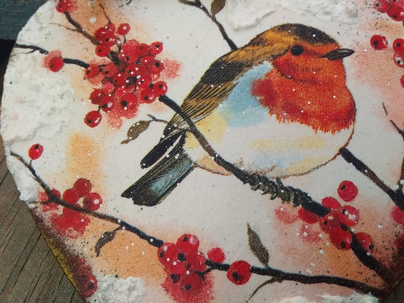 May include: A robin perched on a branch with red berries, painted on a heart-shaped wooden surface with a snowy texture.