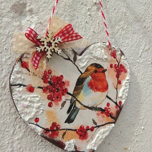May include: A heart-shaped wooden ornament with a robin bird, red berries, and a snowy background. The ornament is decorated with a red and white gingham ribbon and a white snowflake.