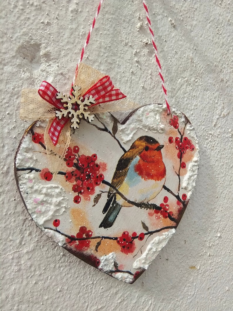May include: A heart-shaped wooden ornament with a robin bird, red berries, and a snowy background. The ornament is decorated with a red and white gingham ribbon and a white snowflake.