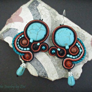 May include: A pair of turquoise and red earrings with a black and white cord design. The earrings feature a turquoise stone with a cracked pattern and a smaller turquoise stone hanging from the bottom.