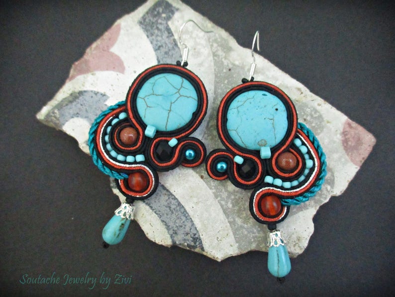 May include: A pair of turquoise and red earrings with a black and white cord design. The earrings feature a turquoise stone with a cracked pattern and a smaller turquoise stone hanging from the bottom.