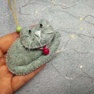 May include: A gray felt cat ornament with a red felt nose and a green bead for an eye. The cat is holding a red felt heart in its paw.