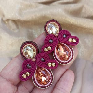 Può includere: Un paio di orecchini in rosa, oro e marrone. Gli orecchini presentano un cristallo a forma di lacrima e dettagli intrecciati intricati.
