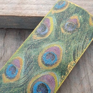 May include: A rectangular wooden bookmark with a peacock feather design. The bookmark is painted in shades of green, blue, purple, and gold. The gold paint is glittery.
